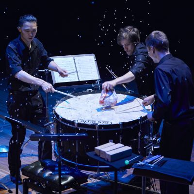 Three percussionists performing around the same drum